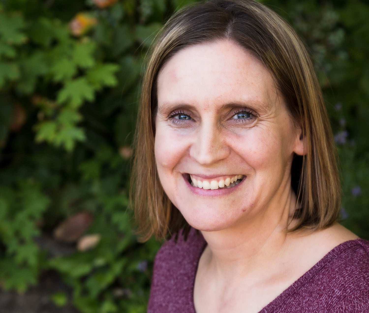 This is a headshot of Tracy Daley, an author for Night Nook Publishing. She has brown hair, blue eyes, and a purple shirt on. She is smiling with a leaf-filled background. 